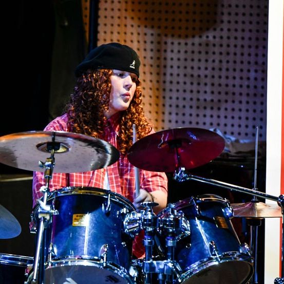 Ein Schlagzeuger mit lockigem Haar spielt auf der Bühne bei einem Festival. Der Schlagzeuger trägt eine schwarze Mütze und ein rotes kariertes Hemd.