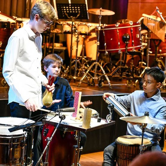 Drei junge Musiker stehen auf der Bühne und spielen Trommeln und eine Gong. Der Junge vorne hält einen Drumstick und eine Gong. Hinter ihm sitzen zwei andere und spielen Trommeln und Becken. Die Bühne ist mit verschiedenen Musikinstrumenten gefüllt.
