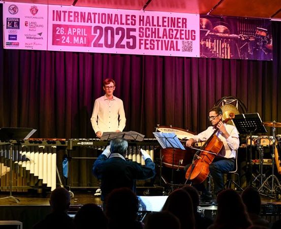 Auf der Bühne findet eine Musikaufführung statt, auf einem Banner steht 'Internationales Halleiner Schlagzeug Festival'. Musiker spielen verschiedene Instrumente, darunter Cello, Schlagzeug und Xylophon. Das Publikum sieht von vorne zu.