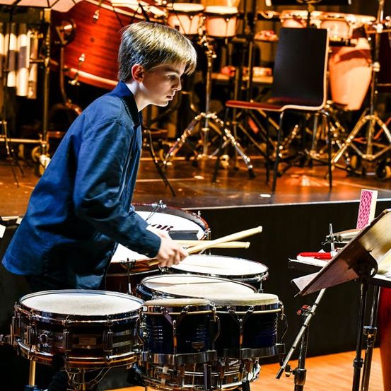 Ein junger Junge spielt auf der Bühne Schlagzeug, umgeben von verschiedenen Musikinstrumenten. Er benutzt Drumsticks und in der Nähe befinden sich Stühle und ein Notenständer.
