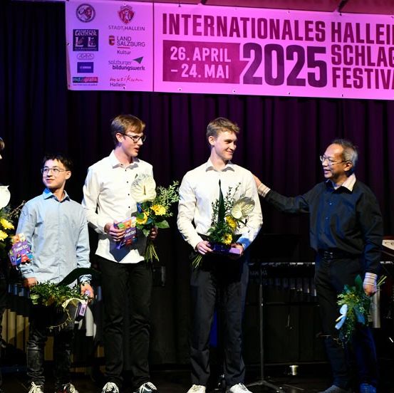 Fünf junge Männer stehen auf einer Bühne, halten Blumen und Trophäen in den Händen, während ein Mann hinter ihnen zusieht. Ein lila Vorhang und ein Schild mit den Worten 'Internationales Halleiner Schlagzeug Festival' sind im Hintergrund zu sehen.