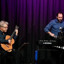 Zwei Musiker treten auf der Bühne auf; einer spielt Gitarre, der andere spielt ein Yamaha-Keyboard mit einer Marimba. Der Hintergrund ist lila.