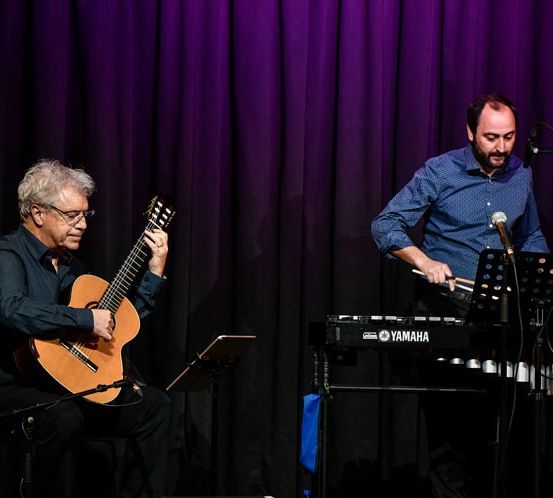 Zwei Musiker treten auf der Bühne auf; einer spielt Gitarre, der andere spielt ein Yamaha-Keyboard mit einer Marimba. Der Hintergrund ist lila.