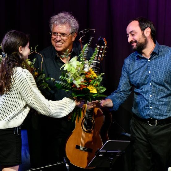 Eine junge Frau überreicht zwei Männern auf der Bühne Blumen, einer hält eine Gitarre und der andere Drumsticks. Sie lächeln und stehen vor einem lila Hintergrund.