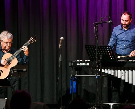 Zwei Musiker treten auf der Bühne auf, mit einem lila Vorhang im Hintergrund. Ein älterer Mann spielt Gitarre, während ein jüngerer Mann Xylophon spielt. Beide konzentrieren sich auf ihre Instrumente.
