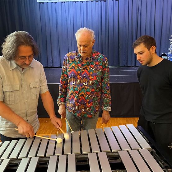 Drei Männer spielen ein Xylophon in einem Raum mit blauem Vorhang und Bühne, einem Schlagzeug und Holzboden.