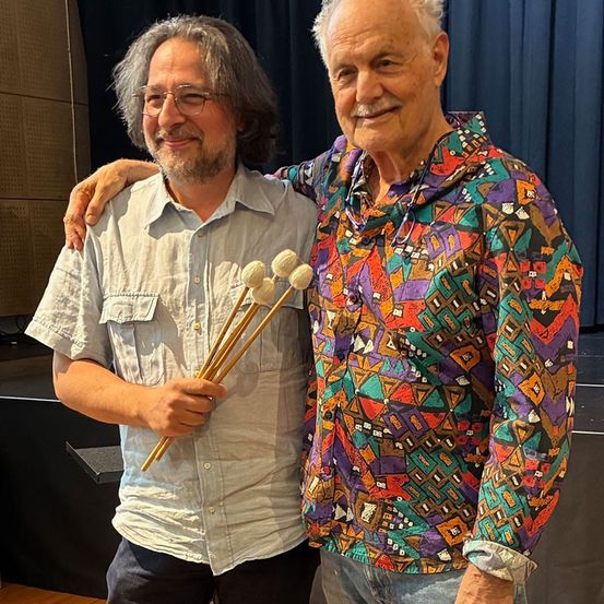 Zwei Männer stehen auf einer Bühne mit einem blauen Vorhang im Hintergrund. Der Mann auf der linken Seite hält Trommelstöcke und der Mann auf der rechten Seite hat seinen Arm um den anderen Mann gelegt.