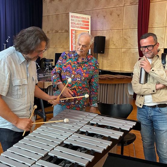 Drei Männer sind in einem Raum mit einem Xylophon. Einer spielt es, ein anderer lächelt und schaut zu, und der dritte verschränkt die Arme. Hinter ihnen steht ein Schild für ein internationales Festival.