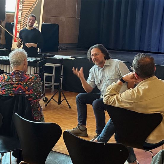 Vier Männer sitzen in Stühlen, dem Bühnenbereich zugewandt. Zwei Männer sitzen, während die anderen beiden auf der Bühne stehen. Ein Mann hält eine Marimba und lächelt.