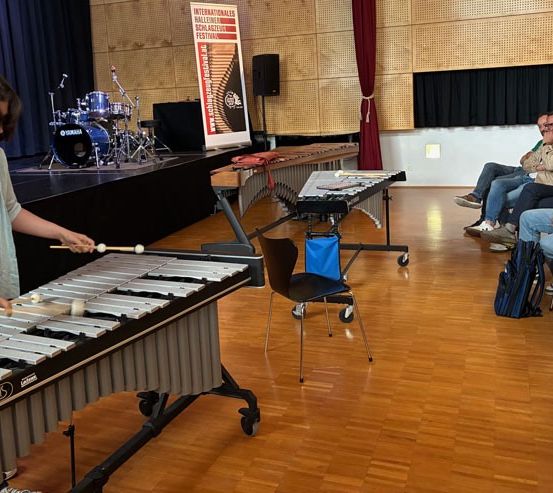 Eine Person spielt ein Xylophon in einem Raum mit Holzböden, während eine Gruppe von Menschen aus Stühlen zusieht. Ein Schlagzeug und ein Banner befinden sich hinter dem Künstler.