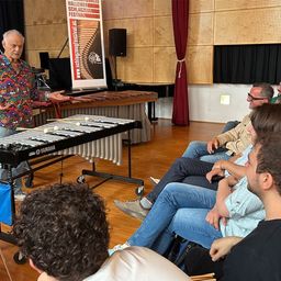 Ein Mann steht vor einem Yamaha-Vibraphon, um ihn herum sitzen mehrere Zuhörer, ein Banner für das Schlageuz-Festival ist im Hintergrund zu sehen.