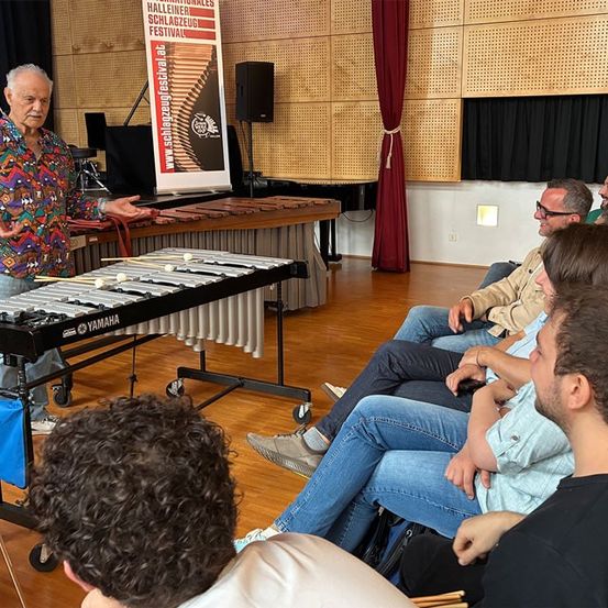 Ein Mann steht vor einem Yamaha-Vibraphon, um ihn herum sitzen mehrere Zuhörer, ein Banner für das Schlageuz-Festival ist im Hintergrund zu sehen.