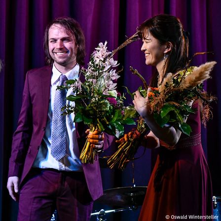 Ein Mann und eine Frau, formell gekleidet, stehen nah beieinander und halten Blumensträuße. Sie lächeln und scheinen bei einer Preisverleihung zu sein. Der Hintergrund ist ein lila Vorhang.