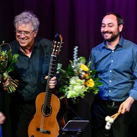 Zwei Männer stehen auf einer Bühne, einer hält eine Gitarre und der andere hält Trommelstöcke. Beide lächeln und zwischen ihnen stehen Blumen. Der Hintergrund ist ein lila Vorhang.