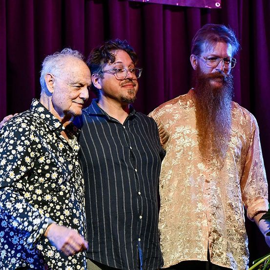 Drei Männer stehen gemeinsam auf einer Bühne, lächeln und posieren für ein Foto. Einer mit Bart und Brille, ein anderer mit einem Blumenhemd und der dritte in einem schwarz-weiß gestreiften Hemd.