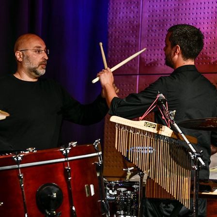 Zwei Männer spielen auf der Bühne Trommeln und Xylophon, einer vor einem großen roten Trommel, der andere mit einer Handglocke in der Hand, vor einem lila Hintergrund.