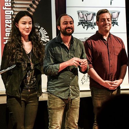 Drei Personen, zwei Männer und eine Frau, stehen auf einer Bühne und lächeln in die Kamera. Sie stehen vor einem Plakat mit Musikinstrumenten und Text.