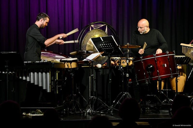 Zwei Männer spielen auf der Bühne Trommeln und andere Instrumente. Sie stehen vor einem Publikum.