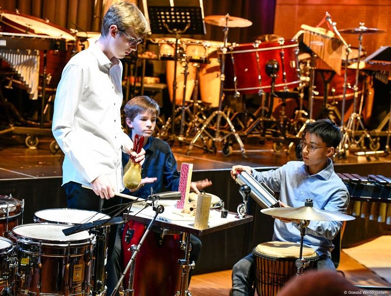 Drei junge Musiker spielen auf der Bühne. Ein Junge hält eine Trompete, ein anderer spielt ein Schlagzeug und ein Dritter spielt ein Drumset. Hinter ihnen sind verschiedene Percussionsinstrumente aufgestellt.