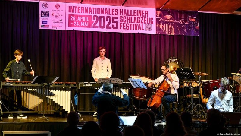 Ein musikalischer Auftritt auf der Bühne mit einem Banner, das 'Internationales Halleiner Schlagzeug Festival 2025' lautet. Ein Cellist, Perkussionisten und ein Violinist spielen vor einem Publikum.