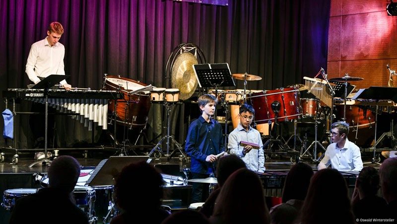 Fünf Musiker treten auf der Bühne auf. Vier von ihnen halten Instrumente, und einer spielt Klavier. Auf der Bühne befinden sich verschiedene Trommeln und Becken. Das Publikum sitzt vor der Bühne.