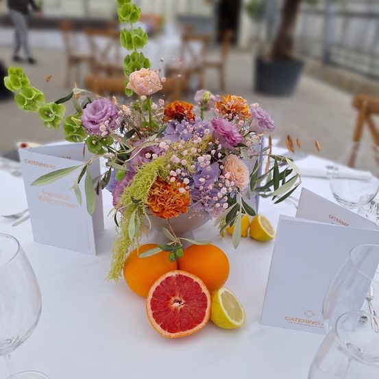Ein Tisch ist für eine Mahlzeit gedeckt mit einem Blumenstrauß, Orangen, Zitronen und Namenskarten. Gläser und Besteck sind ordentlich angeordnet. Ein Topfpflanzen und Stühle sind im Hintergrund.