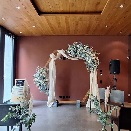Eine Hochzeitseinrichtung mit einem Blumenbogen, der mit weißem Stoff behängt ist, an einer braunen Wand. Stühle und ein Lautsprecher sind aufgestellt. Es gibt einen Fass, ein Tafel und ein Glasfenster.