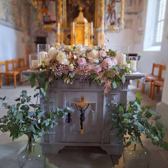 Ein geschmückter Altar mit Kruzifix, rosa und weißen Blumen und Kerzen in einer Kirche. Grüne Pflanzen in Vasen auf beiden Seiten.