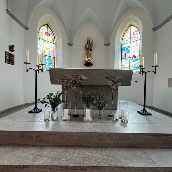 Ein Altar in einer Kirche ist mit Kerzen, einer Statue und bunten Glasfenstern geschmückt. Topfpflanzen und Grünpflanzen sind auf den Stufen des Altars arrangiert.