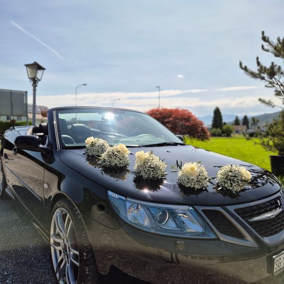 Ein schwarzer Cabrio-Saab ist draußen geparkt, geschmückt mit weißen Blumenarrangements auf seiner Motorhaube und dem Dach, vor einer Kulisse aus Bäumen und Gras.