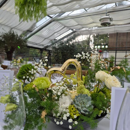 Ein Tisch mit einer großen Herzförmigen Blumendekoration, umgeben von weißen und grünen Blumen. Die Umgebung befindet sich in einem Gewächshaus mit weißen Vorhängen und Grünpflanzen.