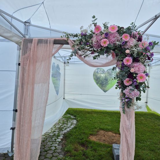 Ein weißes Zelt mit einer floralen Bogendekoration. Der Bogen hat pinke und lila Blumen und Blätter. Das Zelt hat ein herzförmiges Muster auf der Rückseite.