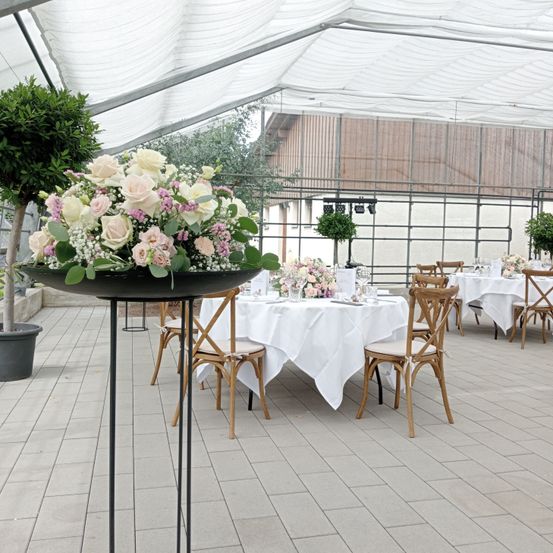 Eine elegante Hochzeitsfeier unter einem weißen Zelt mit weißen Tischdecken und Blumenarrangements in der Mitte. Der Boden ist gefliest, und es gibt Holzstühle und Tische. Eingepflanzte Pflanzen und Bäume ergänzen die Einrichtung.