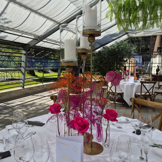 Ein Esstisch mit rosa Rosen und Kerzen in einem Zelt mit Glaswänden. Der Tisch ist mit Gläsern, Gabeln, Messern und Servietten gedeckt. Auf dem Tisch steht ein kleiner weißer Kasten.