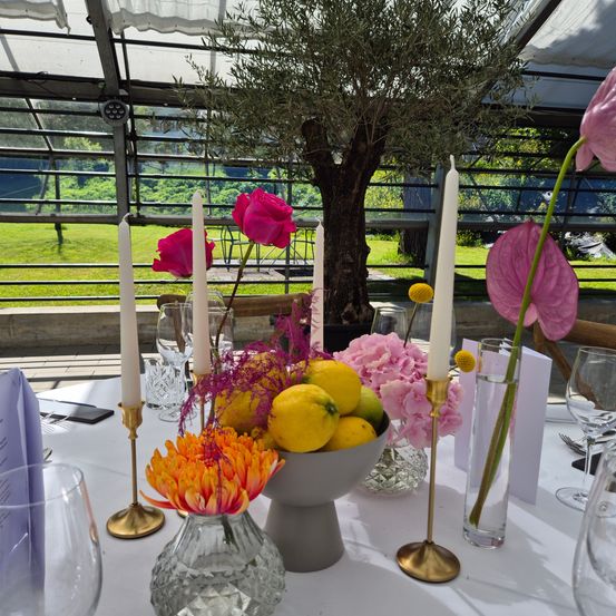 Ein Tisch ist mit Kerzen, Blumen und Zitronen in einem Gewächshaus gedeckt. Ein großer Baum, Grünpflanzen und ein grünes Feld sind durch die Glaswände sichtbar.