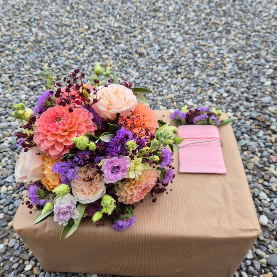 Ein farbenfroher Blumenstrauß mit verschiedenen Rosenfarben, Dahlien und anderen Blumen, auf einer braunen Box mit einer rosa Karte platziert.