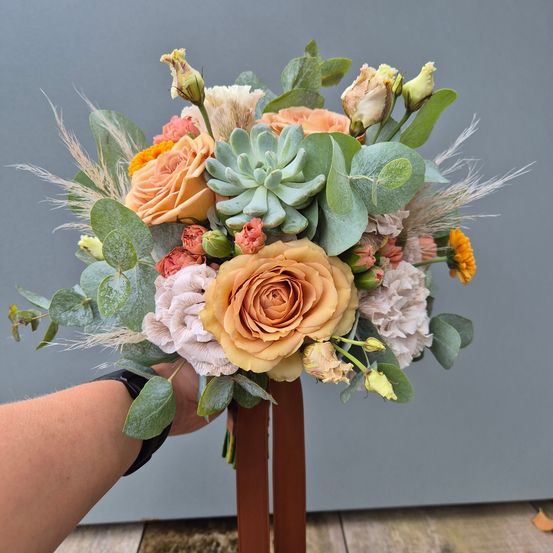 Eine Hand hält eine große Blumenarrangement von Rosen, Sukkulenten und Grünpflanzen, die auf einem braunen Vasen gegen einen grauen Hintergrund ruht.