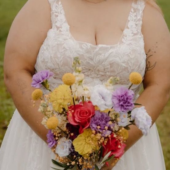 Eine Braut in einem weißen Hochzeitskleid hält einen bunten Blumenstrauß mit roten, gelben und lila Blumen in den Händen.