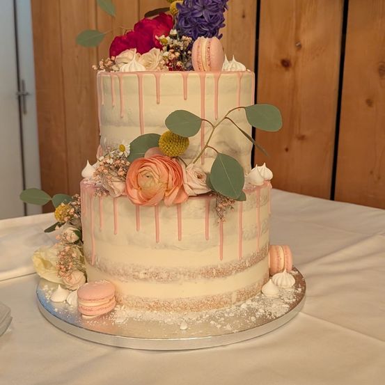Ein dreistöckiger Kuchen mit Blumen, rosa Farbtropfen und Macarons, der auf einem silbernen Ständer auf einem Tisch steht.