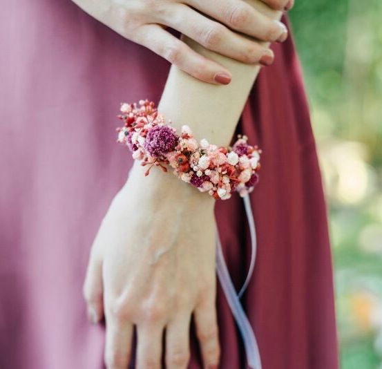 Der Arm einer Person mit einem Blumenarmband. Das Armband besteht aus rosa und weißen Blumen. Die Person trägt ein rosa Kleid.