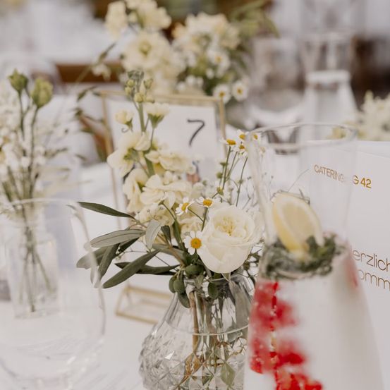 Ein Nahaufnahme einer wunderschön gedeckten Tafel mit weißen Blumen in einer Glasvase, begleitet von anderen Vasen und einem Glas mit Zitronenscheiben. Der Tisch ist Teil einer Veranstaltung oder eines Treffens.