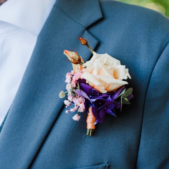Ein blauer Anzug mit einer Ansteckblume, bestehend aus einer weißen Rose, rosa Blumen und grünen und violetten Blättern.