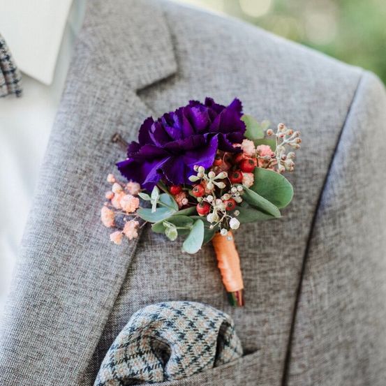 Ein grauer Anzug mit einem grau karierten Taschentuch und einer violetten Blumenbrosche mit rosa und grünen Blumen und Beeren am linken Revers.
