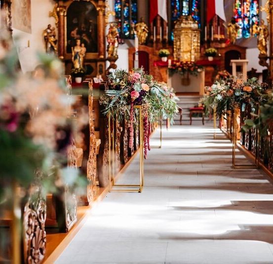 Der Gang einer Kirche ist mit schönen Blumenarrangements geschmückt. Die Kirchenbänke sind mit Blumen und grünen Blättern geschmückt. Der Altar ist mit Blumen und Kerzen geschmückt.