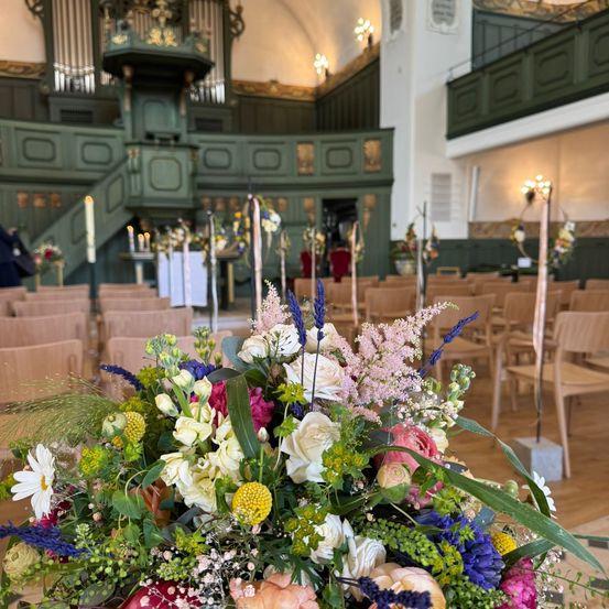 Ein großes Kircheninneres, geschmückt mit einer farbenfrohen Blumenarrangement vorne, umgeben von Reihen hölzerner Stühle und dekorativer Kerzen. Die grüne und weiße Dekoration ergänzt die warmen Holzböden.