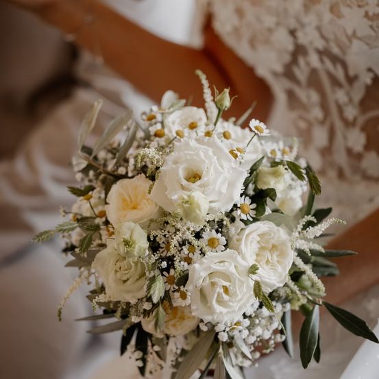 Eine Braut hält einen Strauß weißer Rosen, Babys Atem und Gänseblümchen. Die Blumen sind mit grünen Blättern akzentuiert.
