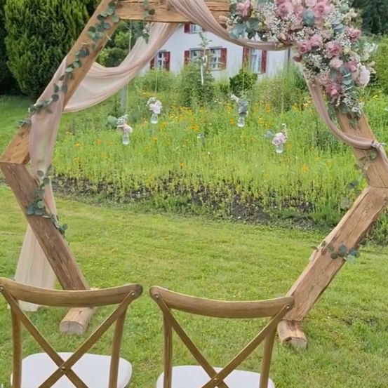 Ein hölzerner sechseckiger Hochzeitsbogen, geschmückt mit rosa Blumen und Grün, steht in einem Feld mit einem Haus im Hintergrund. Zwei Holzstühle mit weißen Sitzen stehen sich gegenüber.