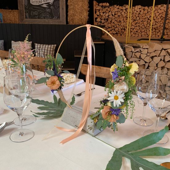 Ein Tisch ist mit einem Blumen-Mittelstück mit einem goldenen Reif, Weingläsern und dekorativen Blättern geschmückt. Im Hintergrund befindet sich ein Kreidetafel-Schild und ein Holzstapel.
