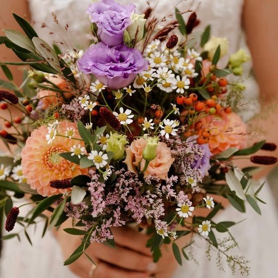 Eine Braut hält einen farbenfrohen Strauß aus gemischten Blumen, darunter lila Rosen, gelbe und weiße Gänseblümchen sowie orange Dahlien, umgeben von üppigen grünen Blättern.