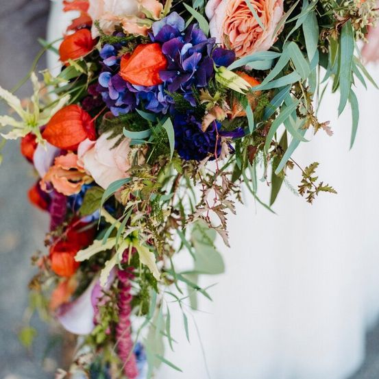 Ein Brautstrauß mit blauen und roten Blumen und grünen Blättern. Die Blumen sind Rosen, Lilien und andere bunte Blumen.